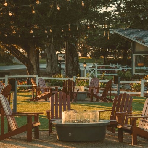 A cozy outdoor fire circle with wooden chairs arranged around a metal fire pit, string lights overhead, and a sunny yard vibe at dusk.