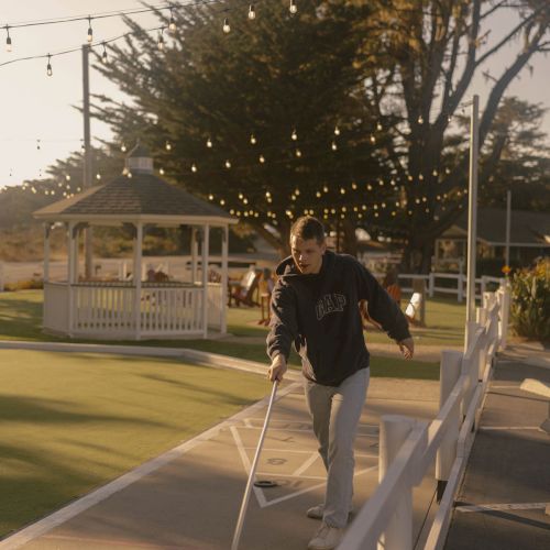 A man uses crutches while walking on a ramp at a sunny park, string lights overhead, a gazebo in the background, and a calm outdoor setting.