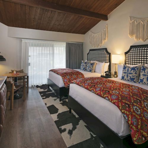 Cozy hotel room with two queen beds, patterned red quilts, blue-and-white pillows, wood ceiling, bedside lamps, desk, TV, and a woven rug.