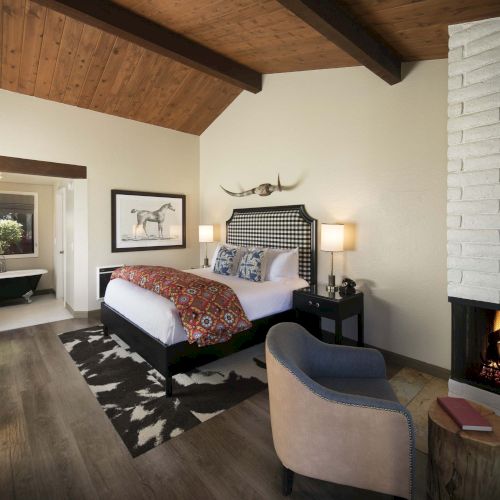 A cozy, rustic bedroom with a large bed, cowhide rug, vaulted wooden ceiling, a fireplace, and a seating area by the fire, in warm tones.