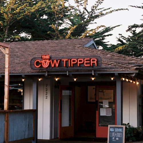 A small restaurant or tavern named &ldquo;The Cow Tipper&rdquo; with a neon sign above the entrance, outdoor chalk board, and string lights.