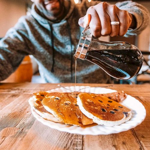 A person in a hoodie pours maple syrup onto a stack of pancakes on a paper plate at a wooden table.