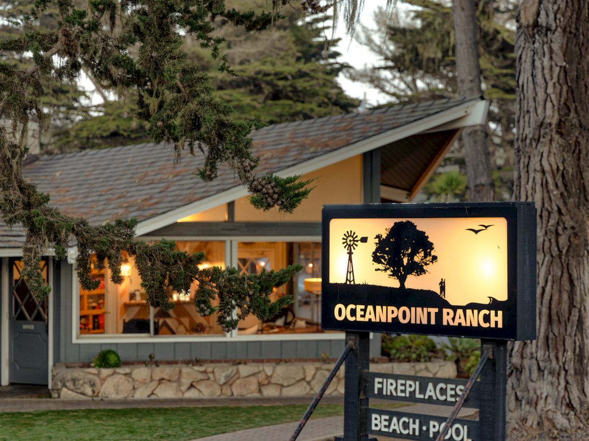 A cozy firehouse-style lodge named Oceanpoint Ranch at sunset, with a lit sign, trees, and a beach pool nearby.