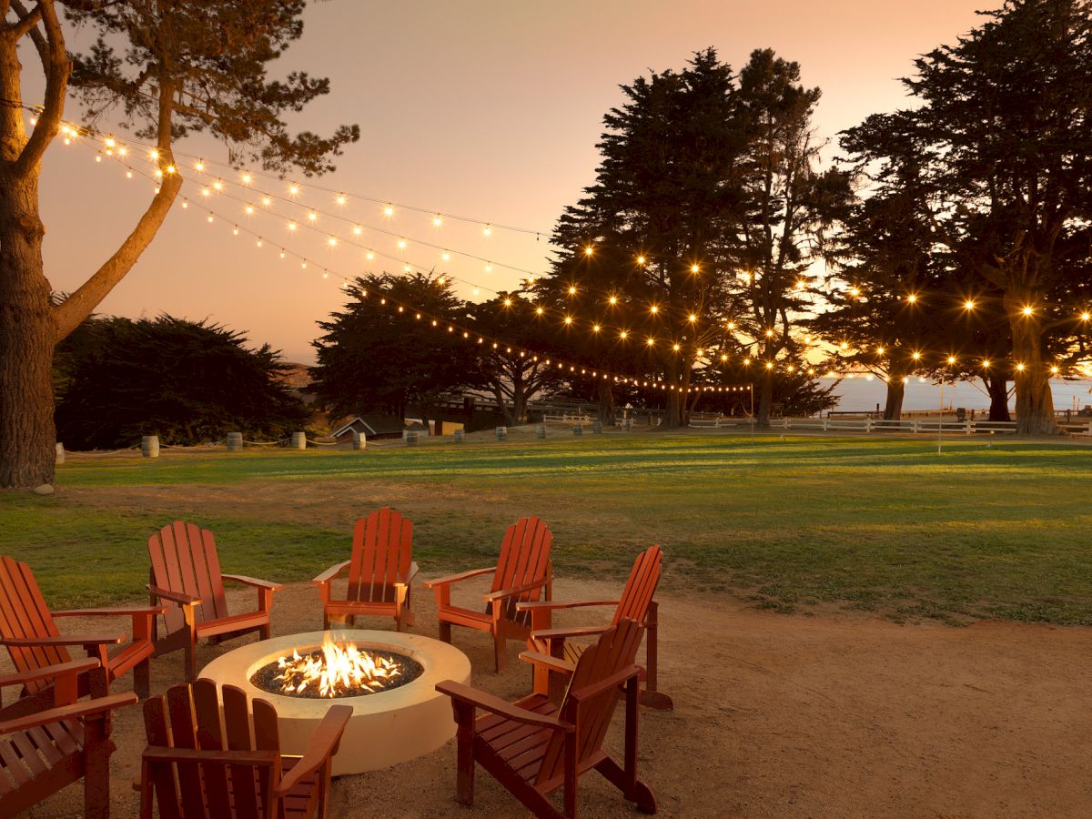 A cozy outdoor gathering with a fire pit, string lights overhead, and wooden chairs arranged in a circle on a grassy lawn at sunset.
