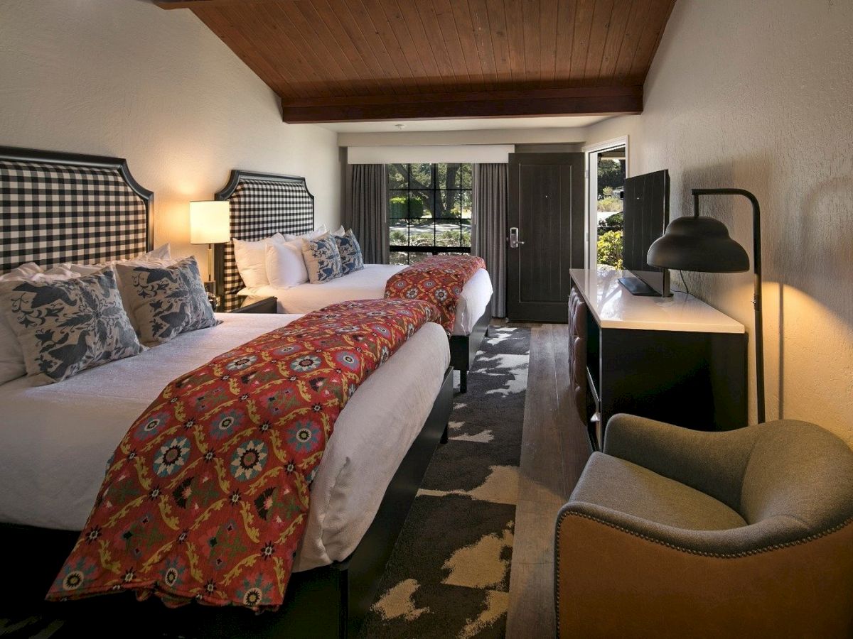 A stylish hotel room with two beds, patterned runners, a checkered headboard, desk, armchair, lamps, and a doorway to a patio.