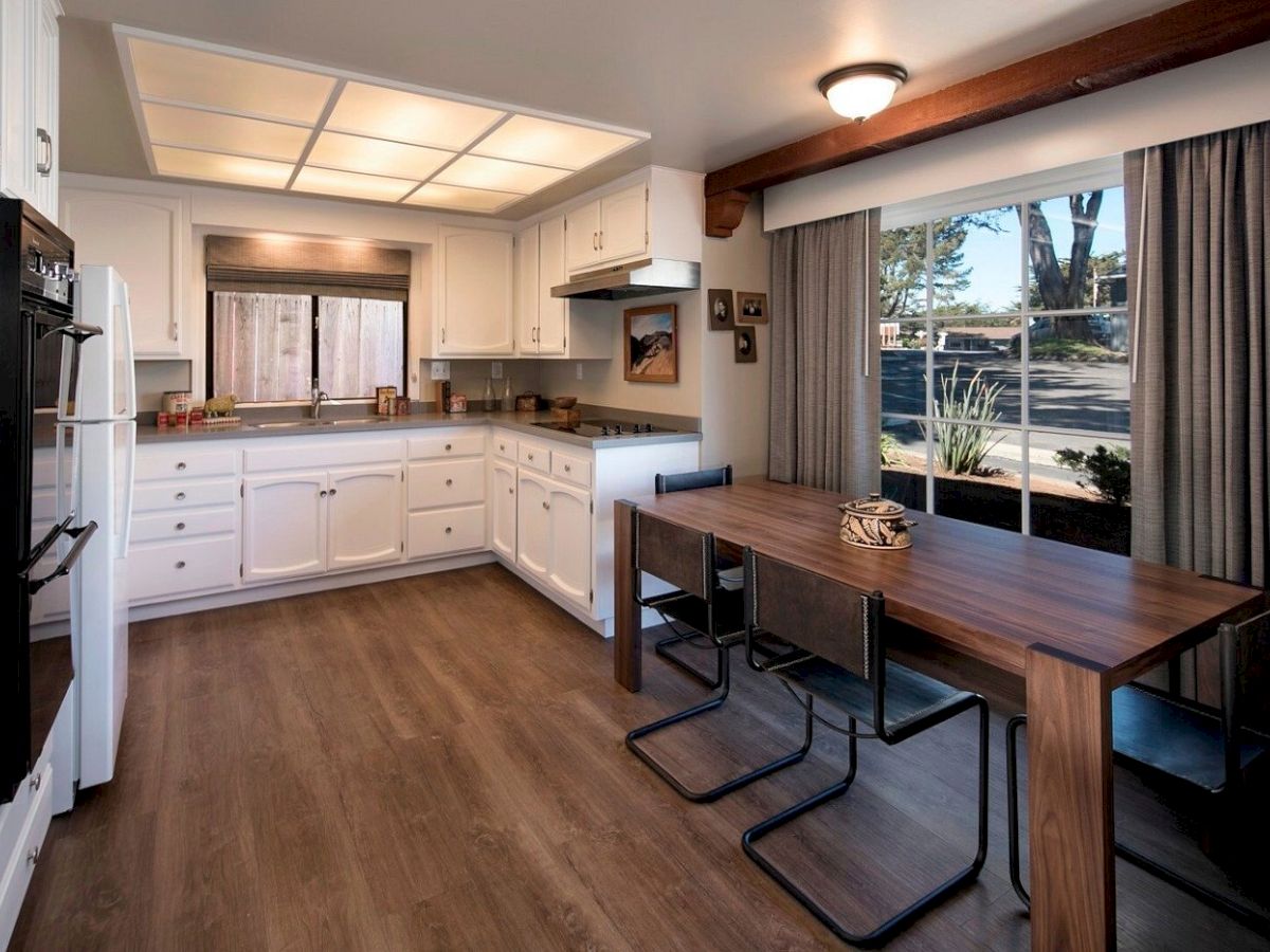 Cozy white U-shaped kitchen with wooden floors, a dining table by a large window, and warm natural light streaming in.