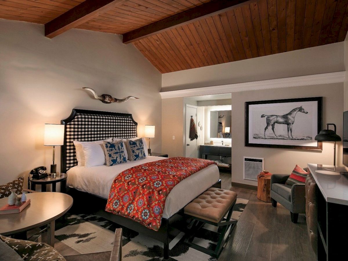A stylish hotel room with a vaulted wooden ceiling, king bed, patterned bedding, two lamps, a tufted bench, and a framed horse illustration.