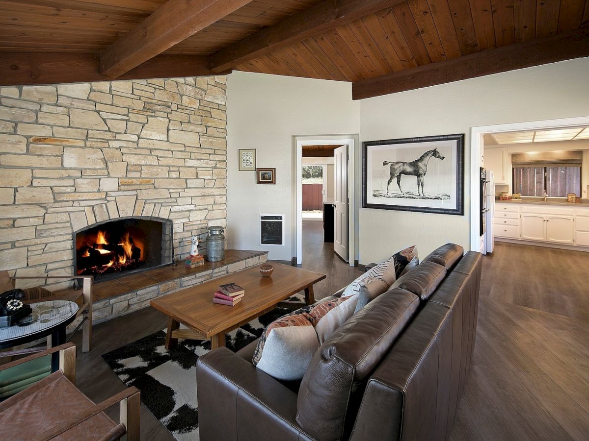 Cozy living room with a stone fireplace, leather sofa, wooden coffee table, rustic decor, and an open kitchen visible in the background.
