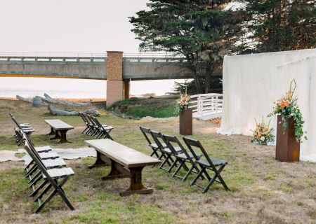 An outdoor wedding setup with a white fabric backdrop, wooden benches and chairs arranged in rows, a small table, flowers, and a nearby bridge over grass.