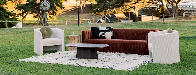 A modern outdoor living area with white sectional sofas, a glass coffee table, and a sunny backyard with trees and a lawn.