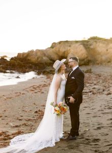 A bride in a white gown and veil with a bouquet stands close to her groom in a suit on a beach, sharing a tender moment at sunset.