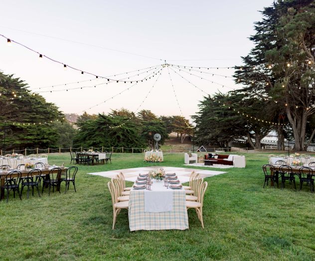 An elegant outdoor setup with long tables, string lights, and a central table on a manicured lawn for a garden party or wedding, under trees.