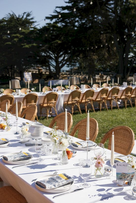 A long outdoor banquet setup with white-tablecloth tables, wicker chairs, floral centerpieces, and elegant place settings under trees, ready for guests.