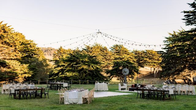 An outdoor event space set up with tables and chairs under string lights, surrounded by trees and rolling hills.