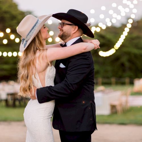 A couple dancing outdoors at dusk, wearing hats, with string lights in the background and a romantic, rustic wedding vibe. End.