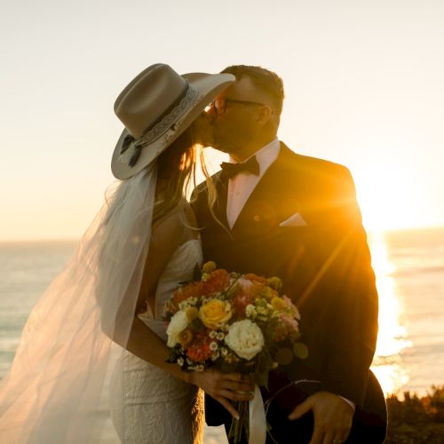 Two silhouettes of a bride and groom kissing at sunset on a beach, the bride holding a bouquet, warm golden light surrounding them.
