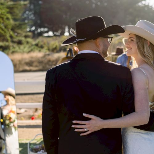 A bride in a white dress and a man in a black suit share a hug at a wedding, with a mirror and other guests in the background, sunny outdoor scene.