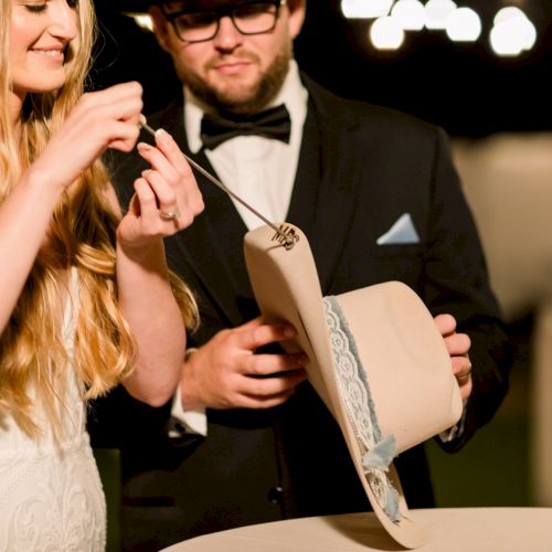 Two people dressed up (a couple) cutting a cake or opening something fancy; the woman in a white dress and the man in a tux, holding a cake knife and a box.