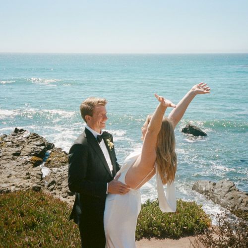 Two people in wedding attire stand on a rocky cliff by the ocean, the bride raising her arms as the groom smiles, sea and sky stretching to the horizon.