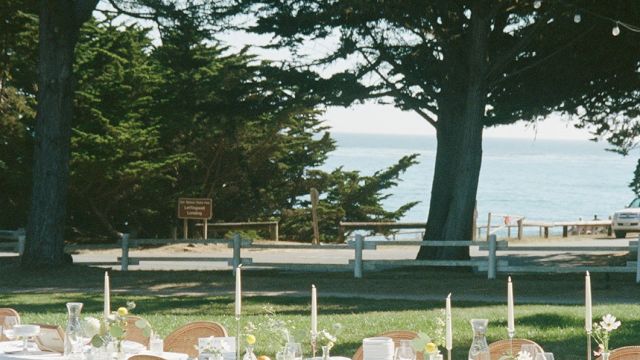 An outdoor event space with a table set for a formal occasion, surrounded by large trees and overlooking the water.