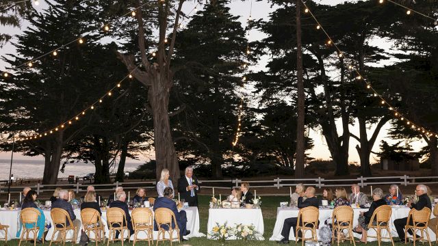 An outdoor wedding reception with tables and chairs set up on the lawn, surrounded by trees and with a view of the ocean in the background.