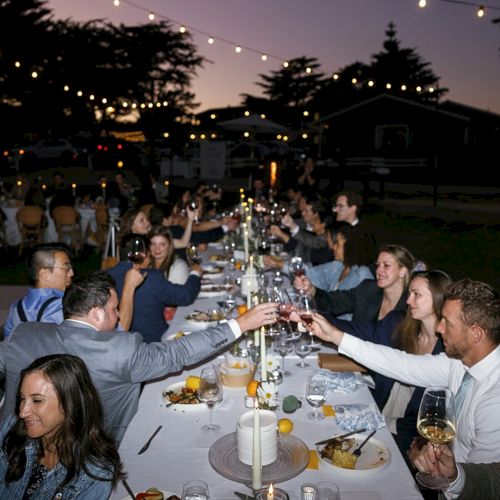 A long outdoor dinner party with string lights, a long table, guests toasting with drinks, and dessert plates in a backyard setting.