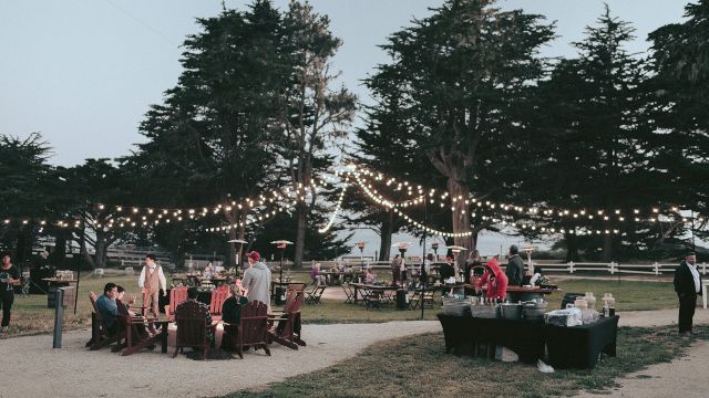 A group of people gathered in an outdoor event space with string lights, tables, and chairs under trees.