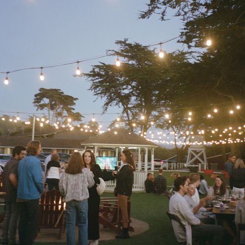 Outdoor evening gathering with string lights, people chatting and sitting at tables under trees and clear sky.