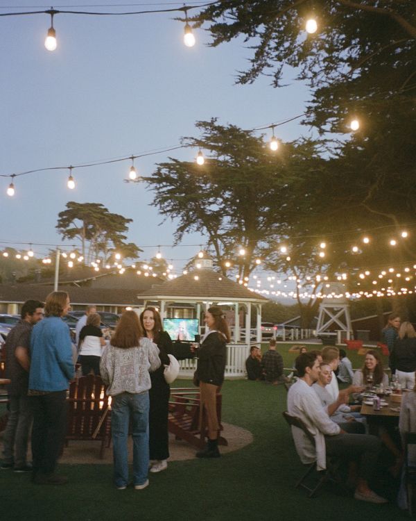 Outdoor evening gathering with string lights, people chatting and sitting at tables under trees and clear sky.
