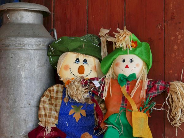Handmade scarecrow dolls in green hats sit by a large metal pot on a red wooden wall, with straw decorations.