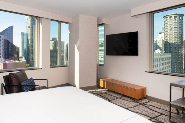 A modern hotel room with a city view, featuring a large bed, a flat-screen TV on the wall, a wooden entertainment center, and a chair by the window.