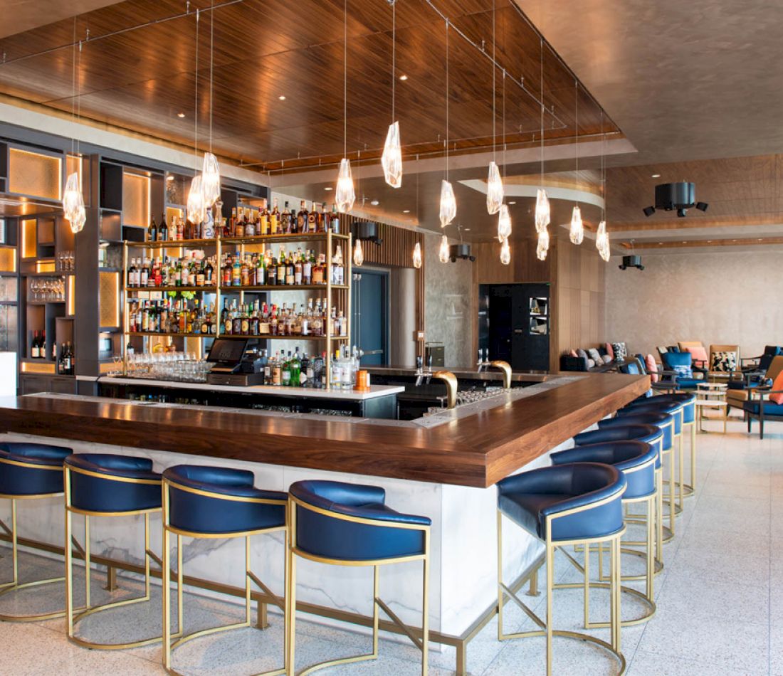 A modern bar with a wooden counter, blue stools, and gold accents, featuring shelves stocked with bottles and glasses.