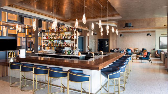 A modern bar with a wooden counter, blue stools with gold accents, and decorative lighting fixtures.
