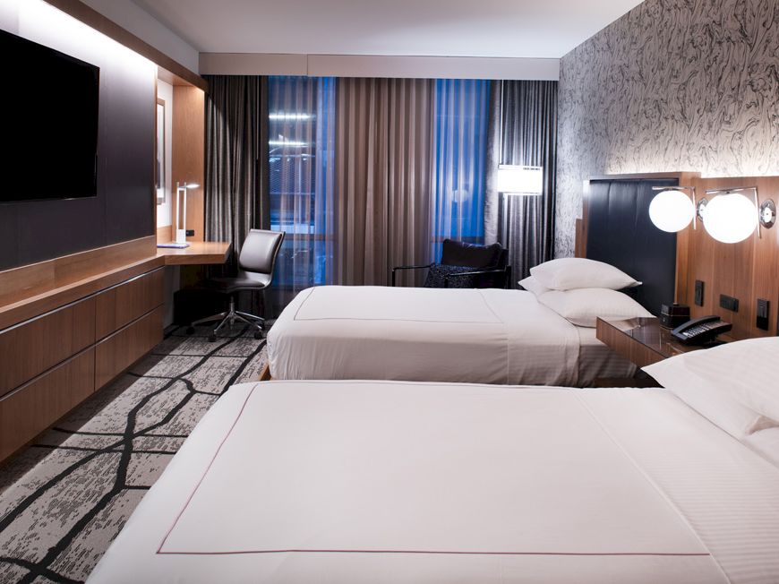 Modern hotel room with two made beds, wall TV, and curtains by the window.