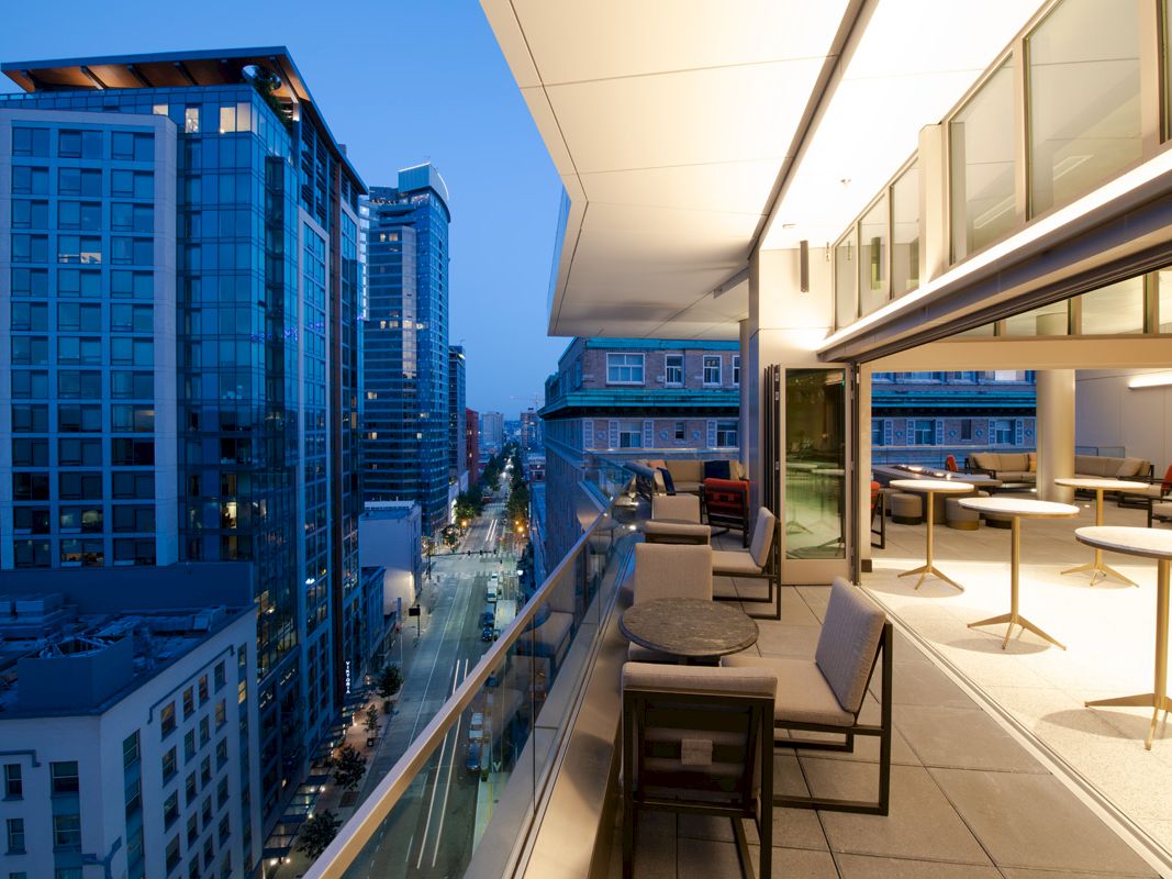 Rooftop terrace with lounge chairs overlooks a downtown street and tall glass buildings under a blue evening sky.