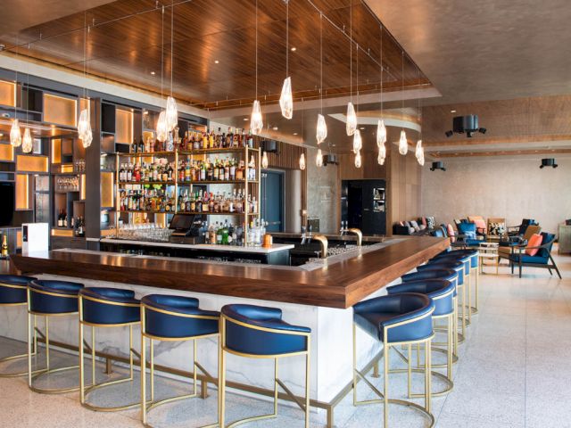 A wide view of a bar with a long wooden counter, blue stools, and gold accents.