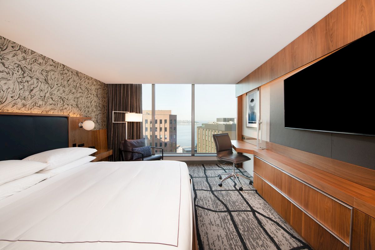 A hotel room with a large bed, desk, TV, and window showing a city view.