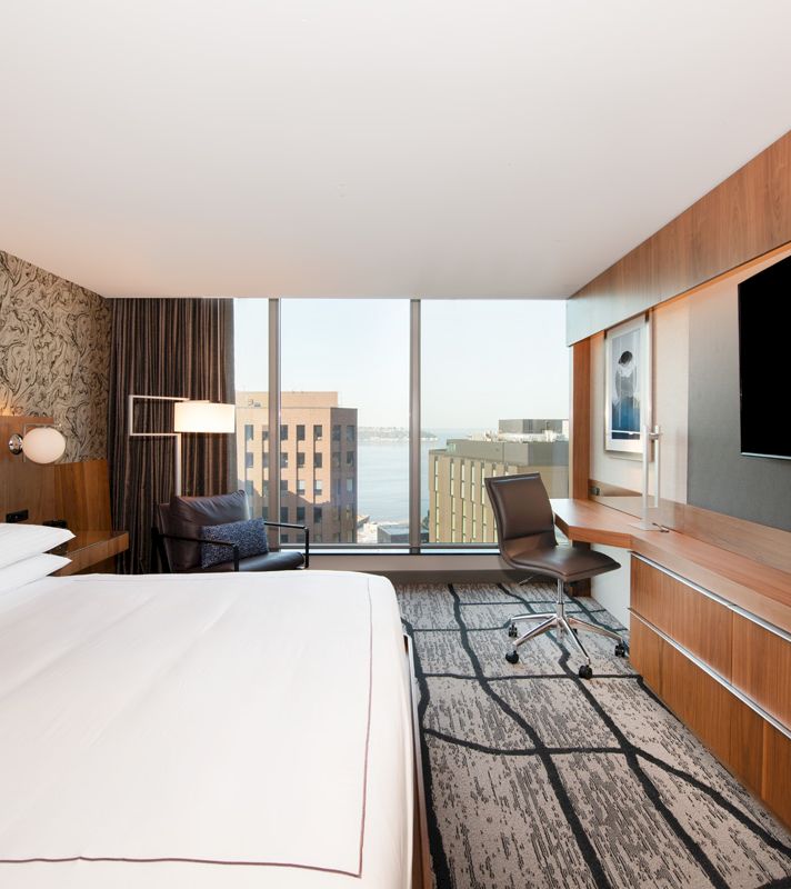 A hotel room with a bed, desk, chair, and TV, and a city view out the window.
